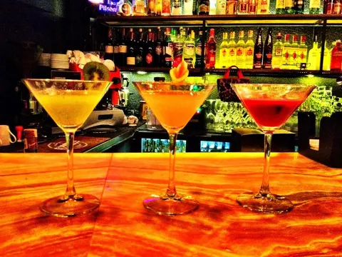 Barista tezgahında üç martini bardağı sıralanmış. Soldan sağa sarı, turuncu ve kırmızı renkli içecekler. Bardakların üzerinde limon ve meyve garnişleri. Arka planda ışıl ışıl şişeler ve bar dekorasyonu. Tezgah turuncu ışıkla aydınlanmış. — KiKi, Sıraselviler 42, Cihangir, İstanbul