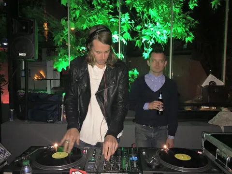 Siyah deri ceket giymiş uzun saçlı DJ, iki platantalı turntable'da müzik çalarken, yanında siyah kazak giymiş bir kişi duruyor. Arkaplan yeşil ışıkla aydınlatılmış yapraklarla süslü. Gece etkinliği atmosferi. — KiKi, Sıraselviler 42, Cihangir, İstanbul