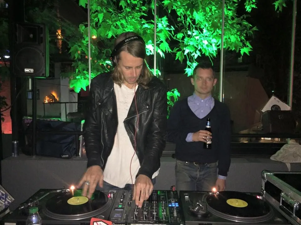 Siyah deri ceket giymiş uzun saçlı DJ, iki platantalı turntable'da müzik çalarken, yanında siyah kazak giymiş bir kişi duruyor. Arkaplan yeşil ışıkla aydınlatılmış yapraklarla süslü. Gece etkinliği atmosferi. — KiKi, Sıraselviler 42, Cihangir, İstanbul
