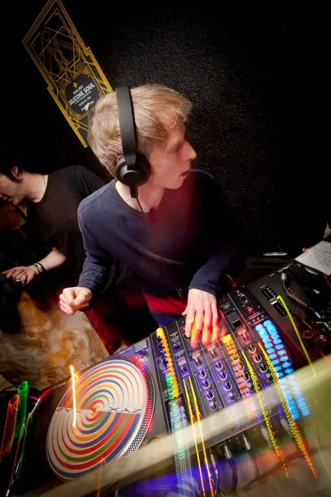 Siyah kulaklıkla DJ kontrol panelinin başında oturan yaşlı müzisyen. Renkli ışıklandırmalı ekranlar ve rengarenk vinil plak görülüyor. Arka planda siyah ışık, önde kırmızı-sarı ışık aydınlatması. — KiKi, Sıraselviler 42, Cihangir, İstanbul