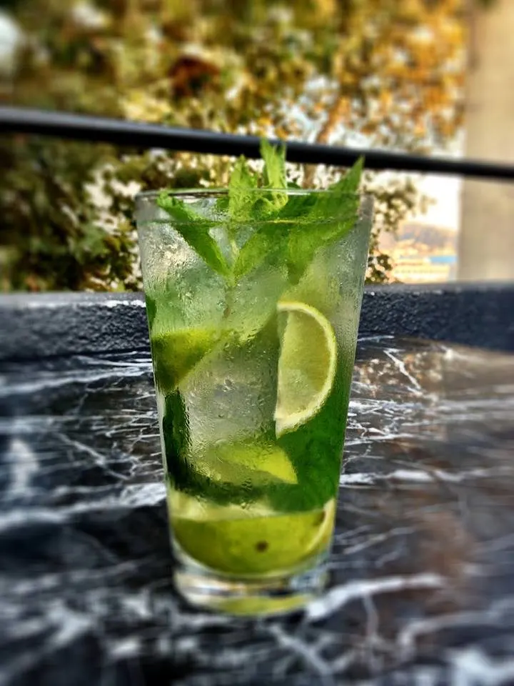 Buz dolu bardakta yeşil mojito kokteyli, taze nane yaprakları ve limon dilimleriyle garnili. Işıl ışıl buharlaşmış camda, bulanık bahçe ortamında güneş ışığı altında. — KiKi, Sıraselviler 42, Cihangir, İstanbul